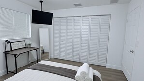 Une chambre à coucher avec un lit, un bureau doté d’une lampe, un téléviseur fixé au mur et une armoire aux portes blanches.