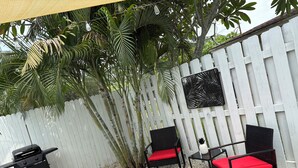 Un espace patio agrémenté d’une clôture blanche, de deux fauteuils en osier noir avec des coussins rouges, d’un barbecue noir et d’un panneau décoratif mural.