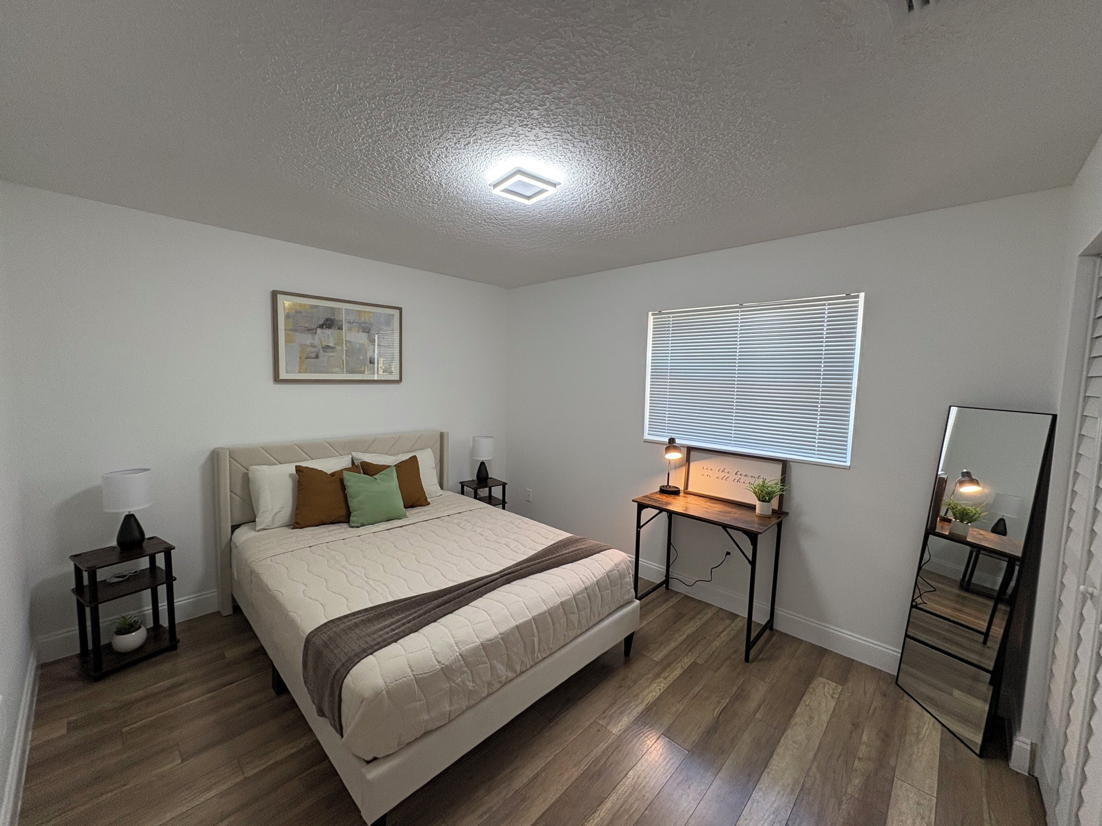 Un dormitorio con cama, mesitas de noche, lámpara de noche, cuadro enmarcado, ventana con persianas y espejo.