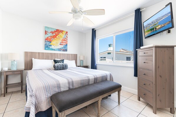 A bedroom with a bed, a bench, a nightstand, a dresser, a ceiling fan, a painting, and a TV.