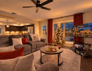 Una sala de estar acogedora con un sofá, una mesa de centro con aperitivos y vino, un árbol de Navidad y un televisor que muestra una escena navideña.