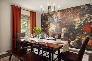 Un comedor con una mesa de madera dispuesta para cuatro personas, una araña y un mural floral en la pared.