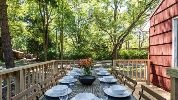 An outdoor dining area with a long table set for six, surrounded by wooden railings, with trees in the background.