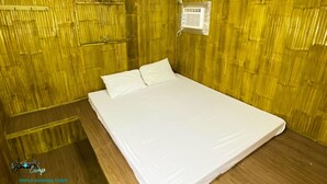 A triple sharing cabin with a wooden wall, a bed with white bedding, and a small air conditioning unit. - Spark Camp - Baler (Baler)