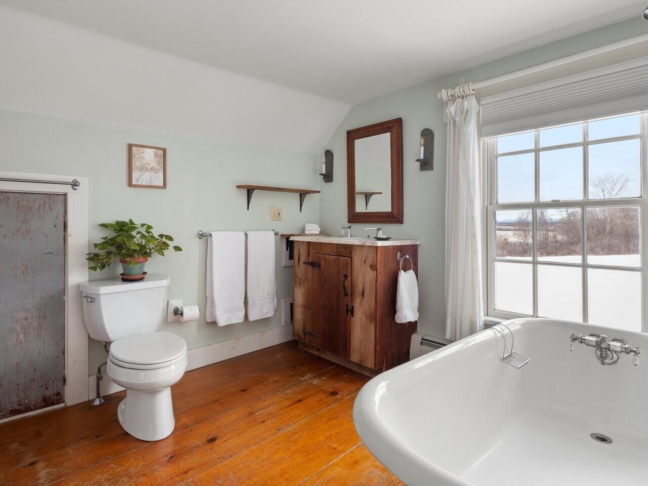 A bathroom with a wooden floor, white fixtures, a large window, and a view of the outdoors.
