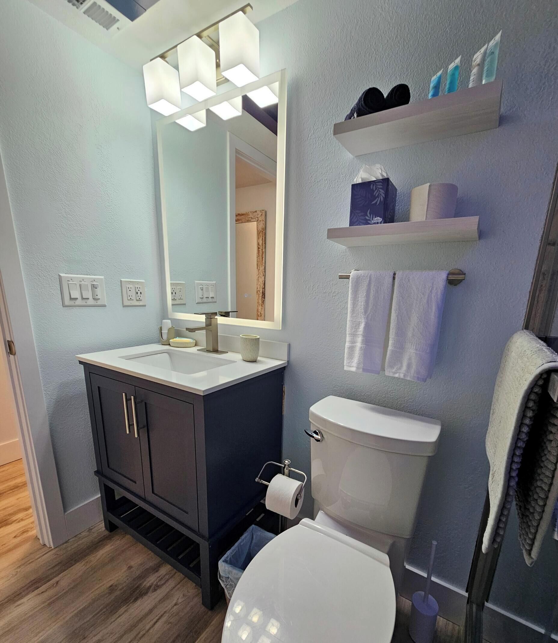 A modern bathroom with a white toilet, dark cabinets, a mirror with integrated lighting, and a shower area.