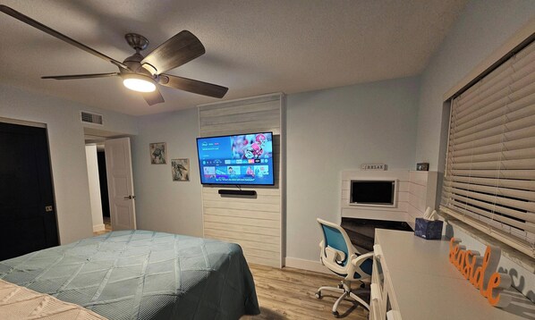 Un dormitorio con una cama, un escritorio con computadora, un ventilador de techo y un televisor en la pared.