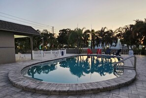 Una piscina con un cartel que indica un límite de profundidad de 6 pies, rodeada por un pavimento de ladrillo, palmeras y una zona cubierta con sillas rojas.