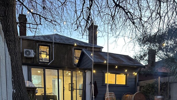 A modern house with a fire pit in the foreground, a dining area with a table and chairs, and a large window overlooking the backyard. - Atelier’s Den-Wood Fire Hot Tub, Centre Of Town (Beechworth)