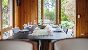 A dining table set with plates, glasses, and a bowl, surrounded by chairs, in a room with wooden walls and a view of greenery outside. - Spacious | Pet Friendly| Near Boat Ramp and Water (Clifton Springs)