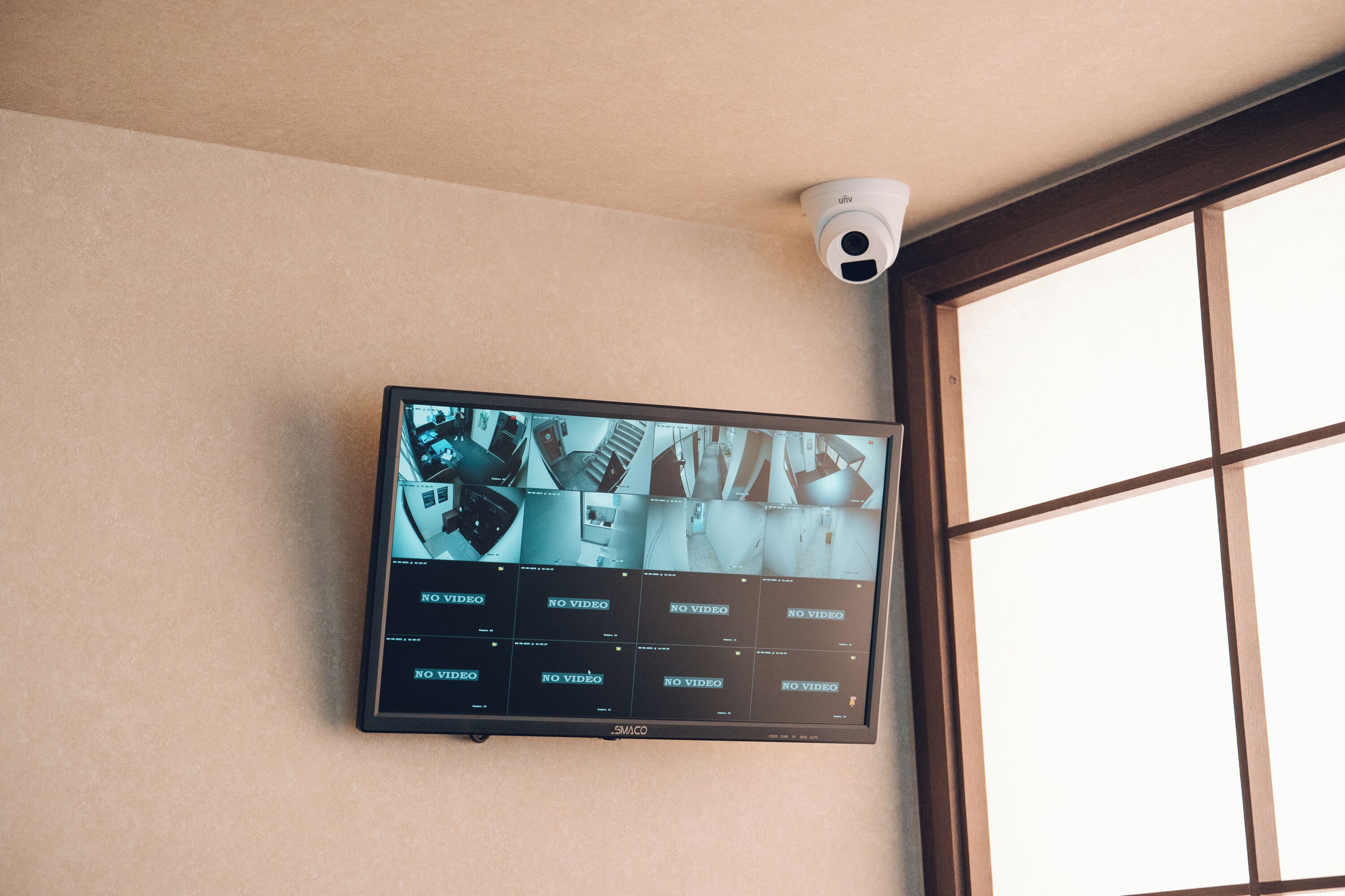 A security camera mounted on the wall above a flat-screen TV displaying multiple camera feeds with "NO VIDEO" messages.