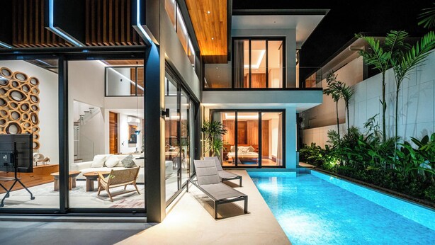 Una casa moderna con un área de estar con paredes de vidrio, una piscina y un balcón con plantas.