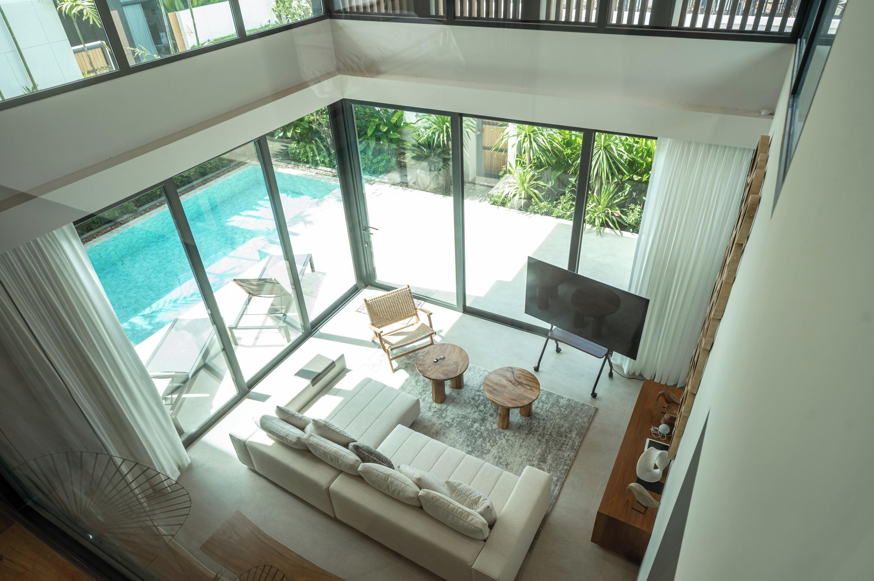 A spacious living area with a large sofa, two wooden side tables, and a flat-screen TV. A glass door leads to an outdoor swimming pool area with a chair and a table.