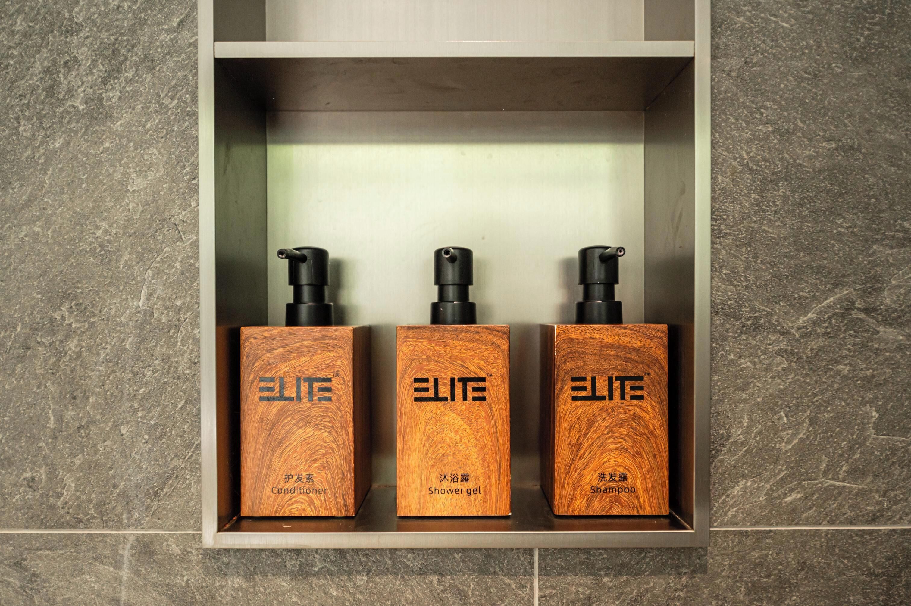 Three wooden bottles with black pumps, labeled 'ELITE', on a shelf against a tiled wall.