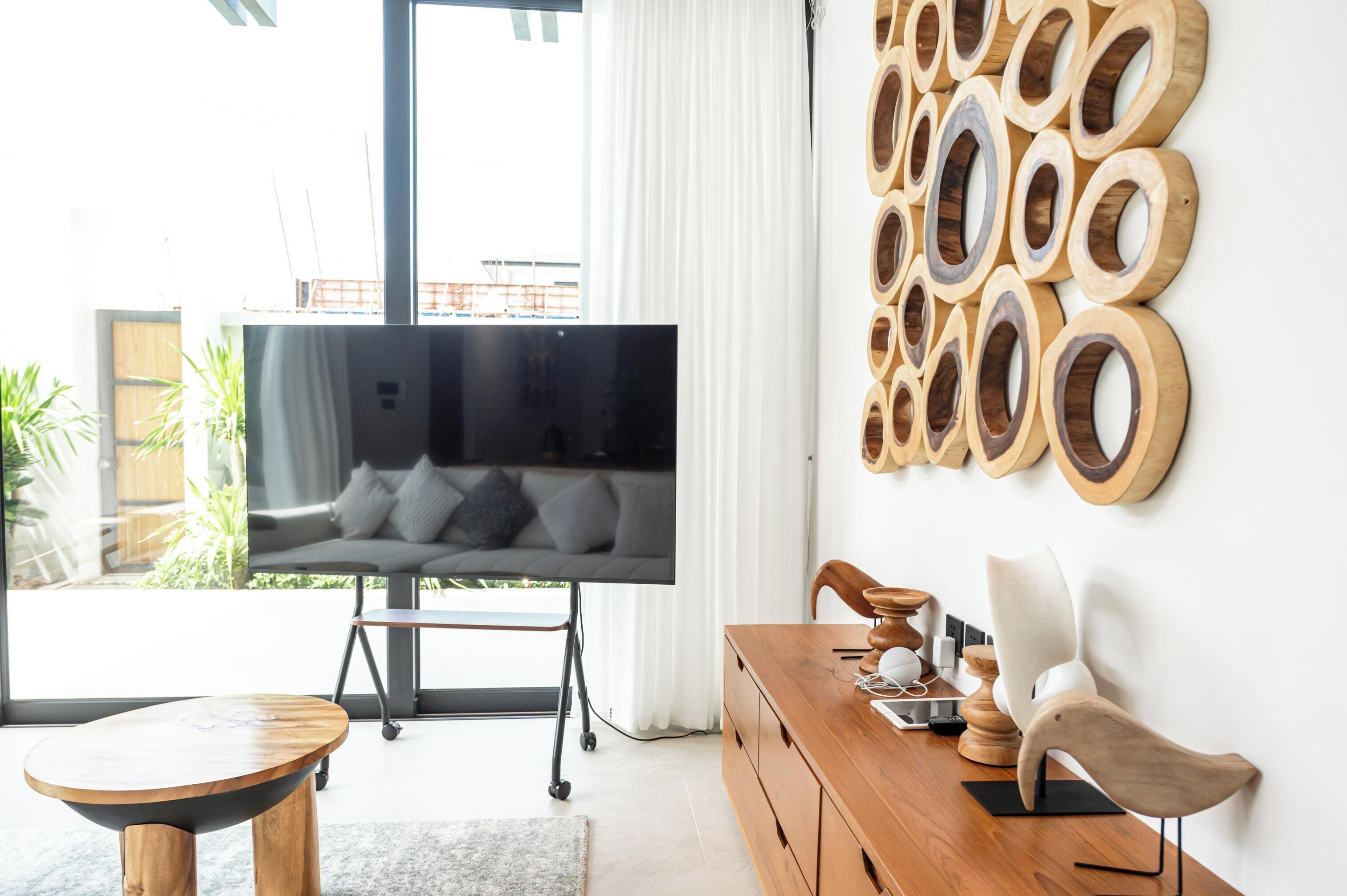 A modern living room with a flat-screen TV, a wooden dining table, and a wall-mounted wooden art piece.