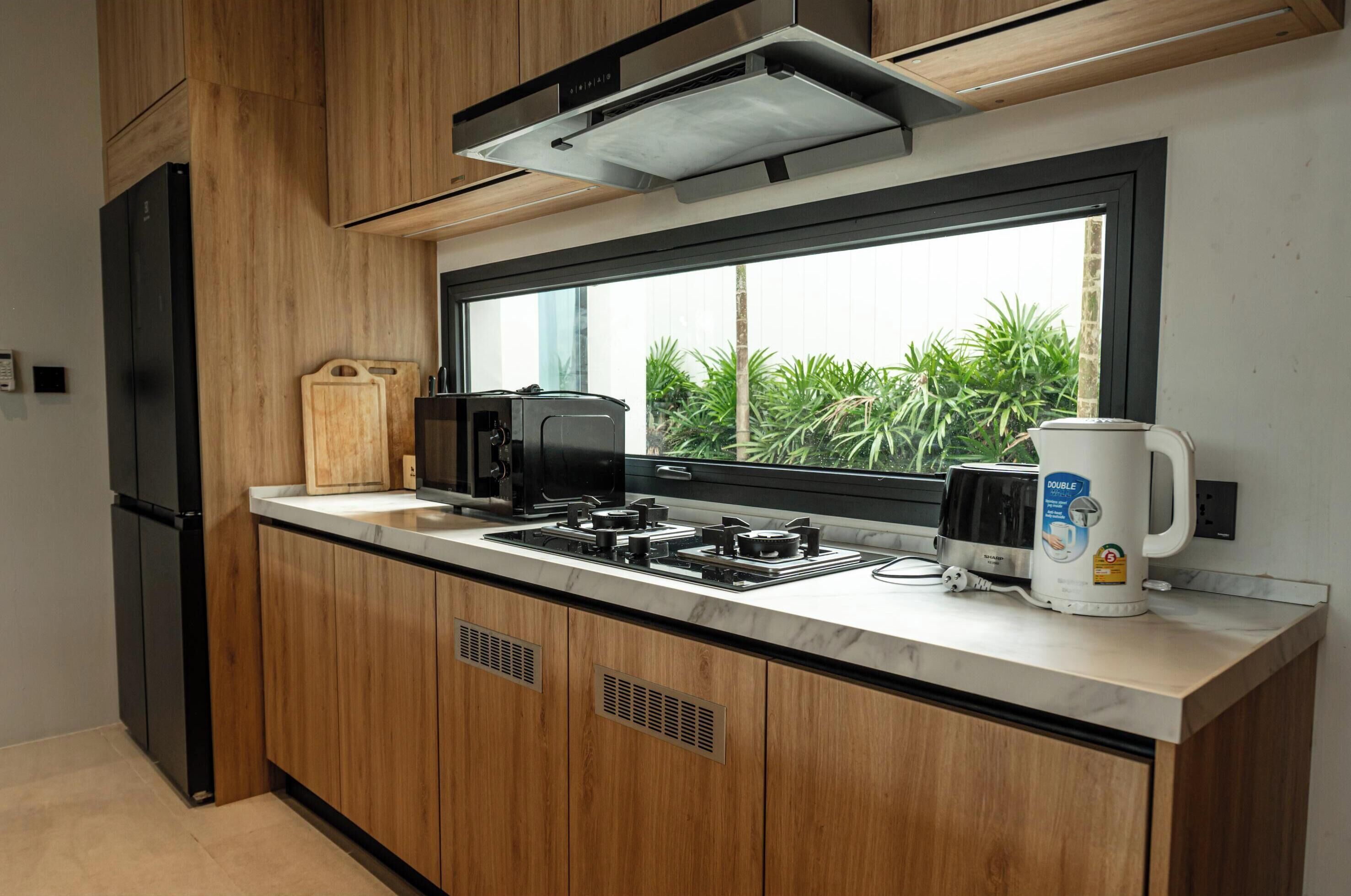 A modern kitchen with wooden cabinets, a built-in stove, a coffee maker, and a window overlooking greenery.