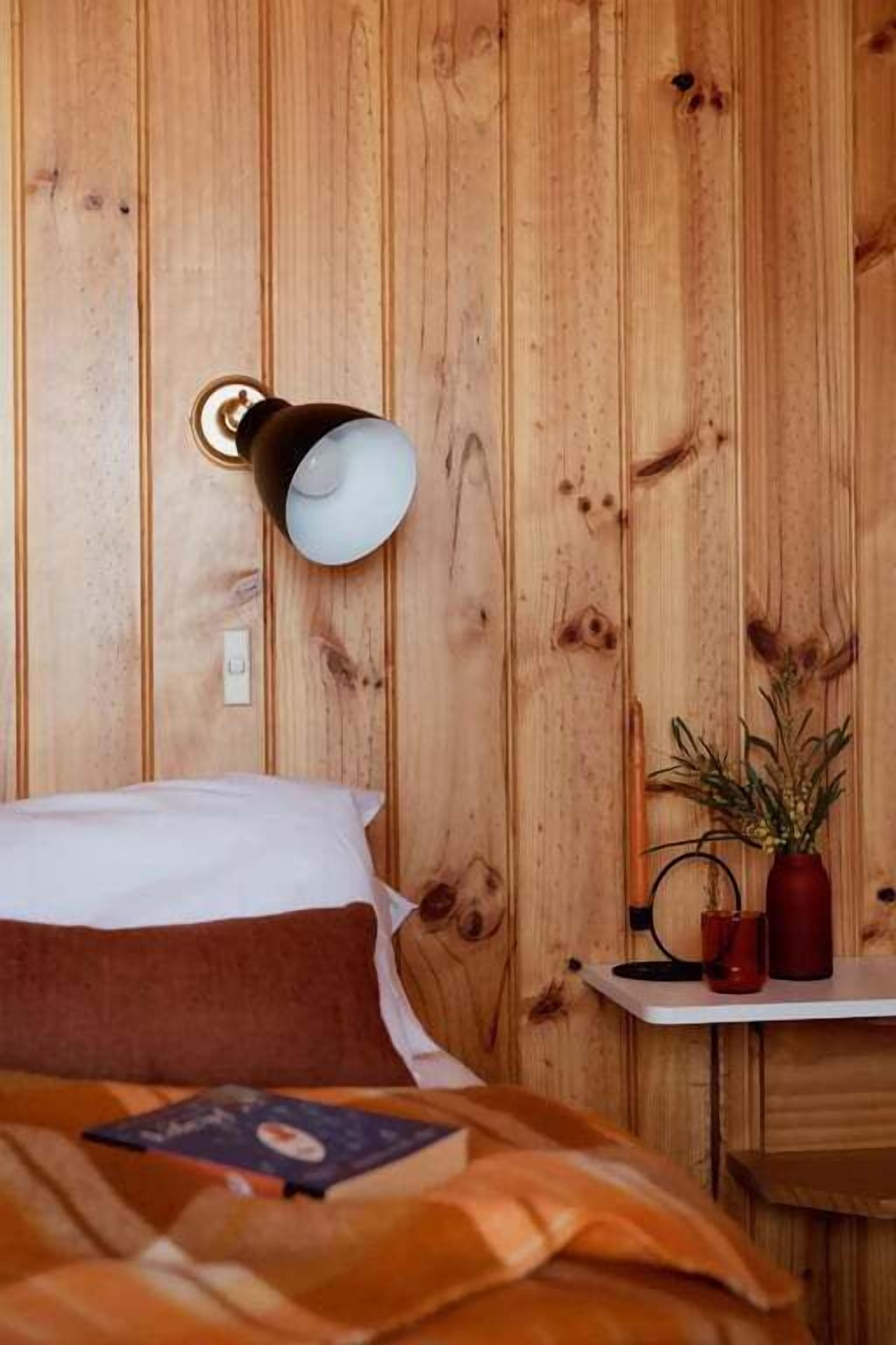 Ein Zimmer mit Holzwandverkleidung, einem Bett, einer Wandlampe und einem kleinen Tisch mit einer Pflanze und zwei roten Tassen.
