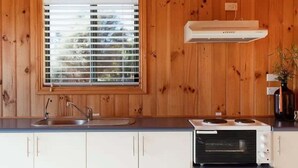 A kitchen with wooden paneling, a window with blinds, a sink, a stove, and a wall-mounted air vent. - Panorama Cabin - a cosy shack and an epic view (A) (Bicheno)