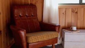 A wooden-paneled room with a brown leather armchair and a white dresser with a cup and book on top. - Panorama Cabin - a cosy shack and an epic view (A) (Bicheno)