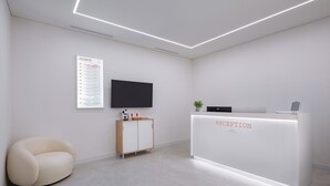 Un mostrador de recepción con un cartel, un televisor de pantalla plana, un mueble de madera, una silla acolchada y una planta.