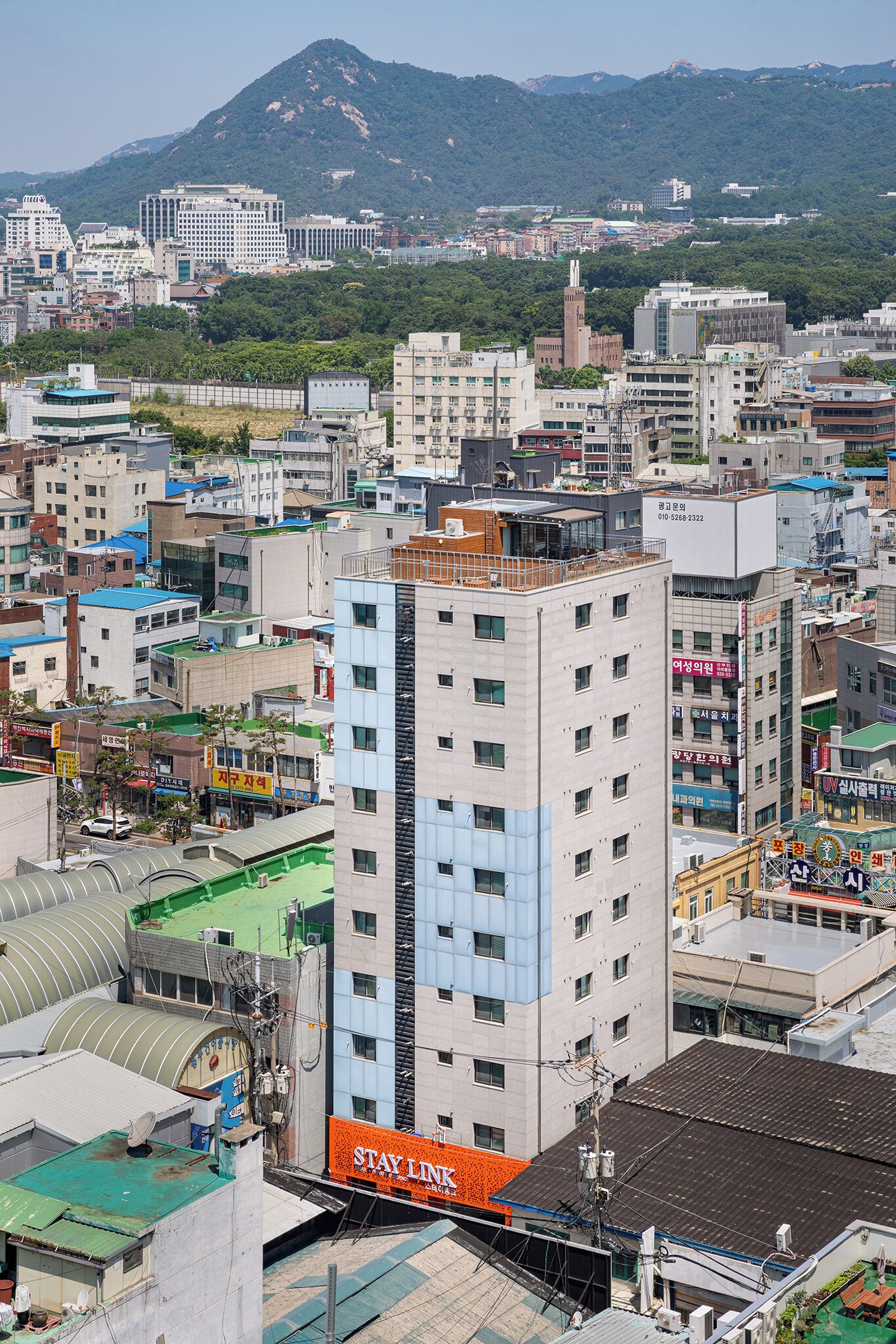 A multi-story building with a prominent 'Stay Link' sign, surrounded by other buildings and a mountainous backdrop.