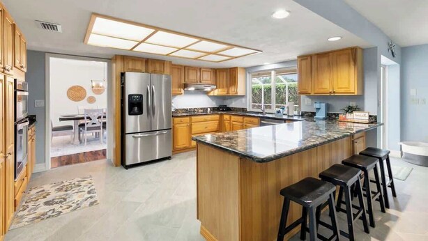 A modern kitchen with wooden cabinets, a large island with bar stools, stainless steel appliances, and a view to the dining area. - Sunny Marco Home with Heated Pool Near Beaches (Marco Island)