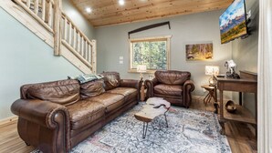 A spacious living room with a wooden staircase, leather sofas, a coffee table, and a large window with a view of trees.
