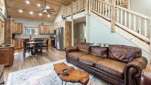 A spacious living area with a brown leather sofa, wooden dining table, and chairs. A wooden coffee table is in the center. The room has wooden floors, a high ceiling with exposed beams, and a staircase with a wooden railing.