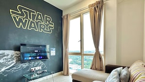 Una sala de estar moderna con una pared temática de Star Wars, un televisor de pantalla plana, una mesa de centro de vidrio, un sofá y vistas de un paisaje urbano a través de amplias ventanas.