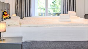 Una cama bien hecha con sábanas blancas, un peluche y una lámpara de noche en una habitación con ventanas amplias y cortinas grises.