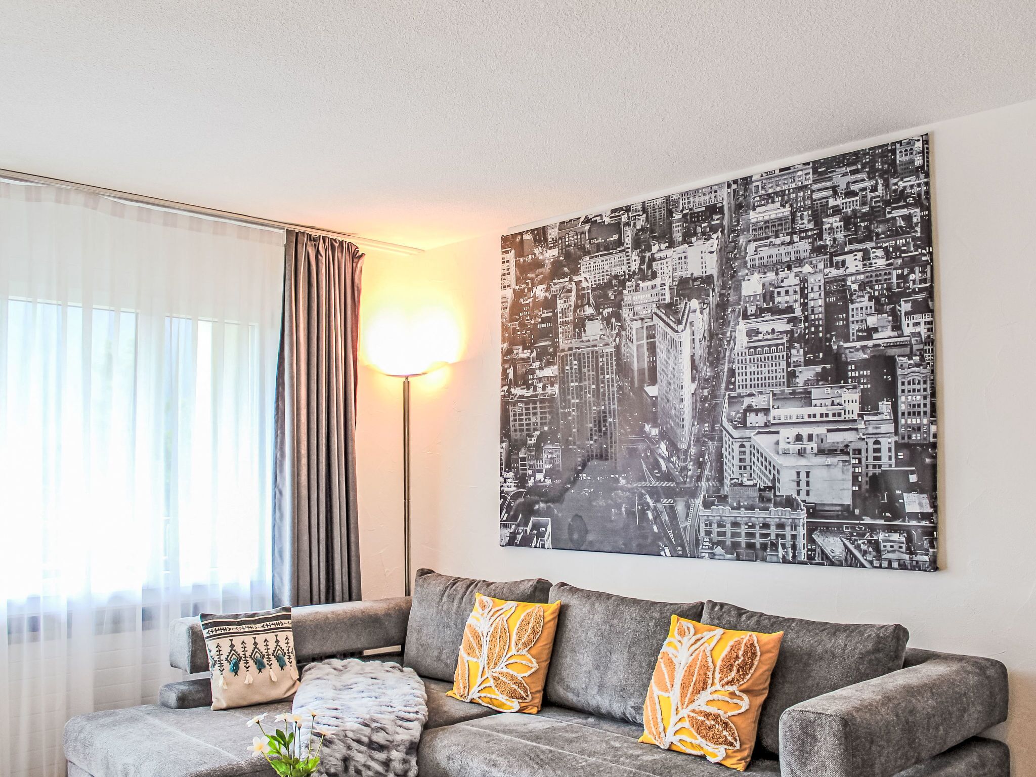 Uma sala de estar moderna com um sofá cinzento, almofadas decorativas, um abajur e uma pintura com vista para a cidade na parede.