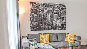 Una sala de estar con un sofá gris, dos almohadas amarillas, una almohada estampada, una pequeña mesa de centro de madera y un cuadro con un paisaje urbano.