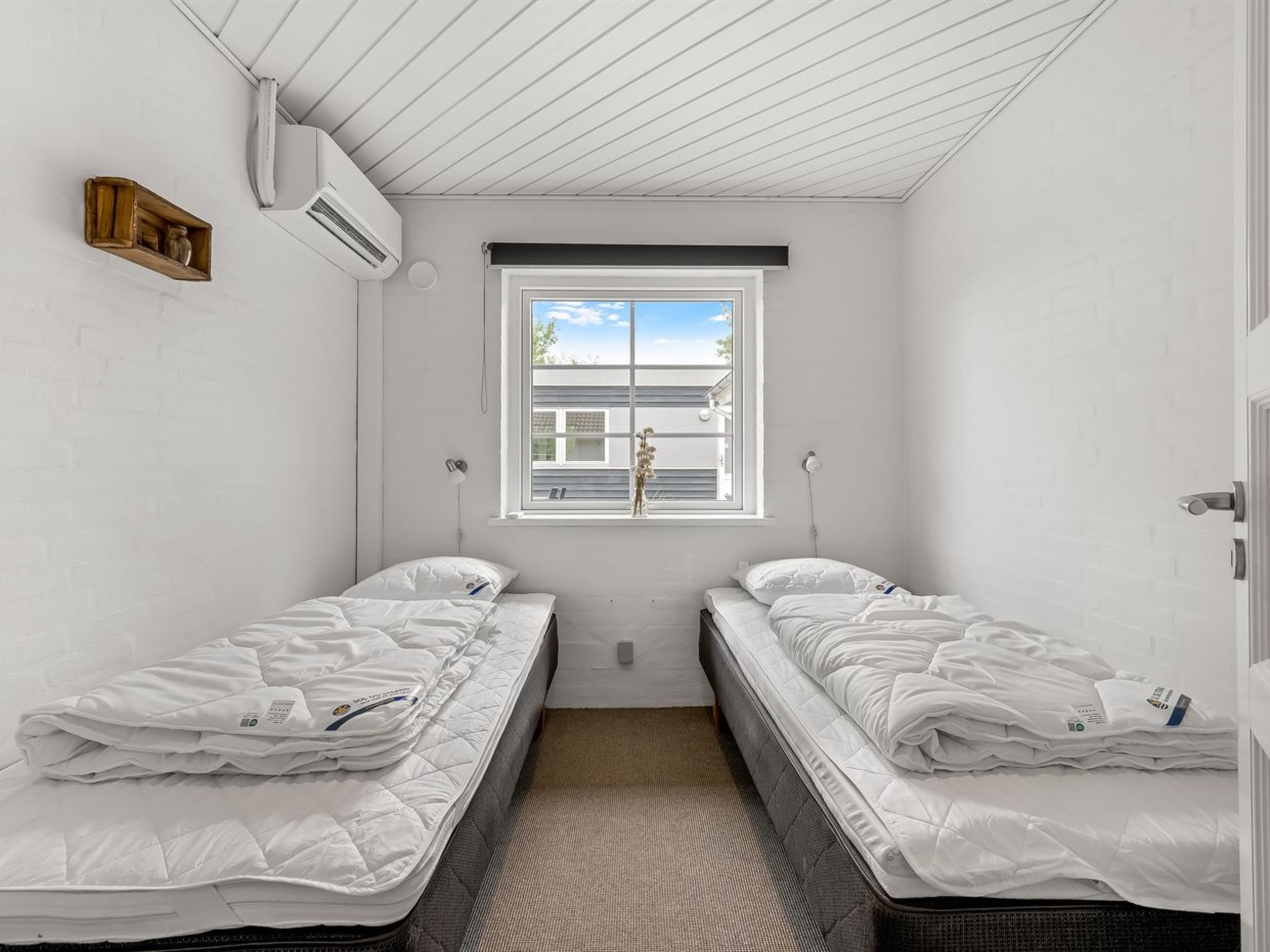 A small, clean room with two single beds, a window with blinds, an air conditioning unit, and a small shelf.