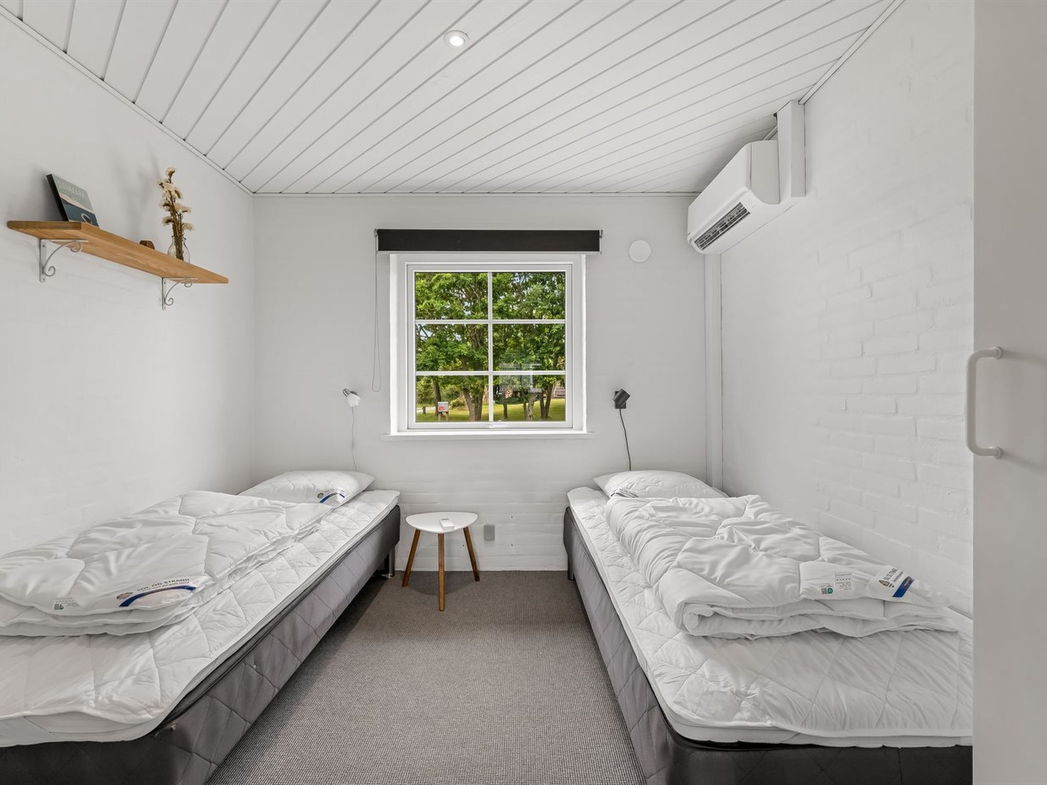 Ein kleines, modernes Zimmer mit zwei Einzelbetten, einem kleinen Tisch, einem Fenster mit Blick auf Grünflächen und einer Wand aus weißen Ziegelsteinen.