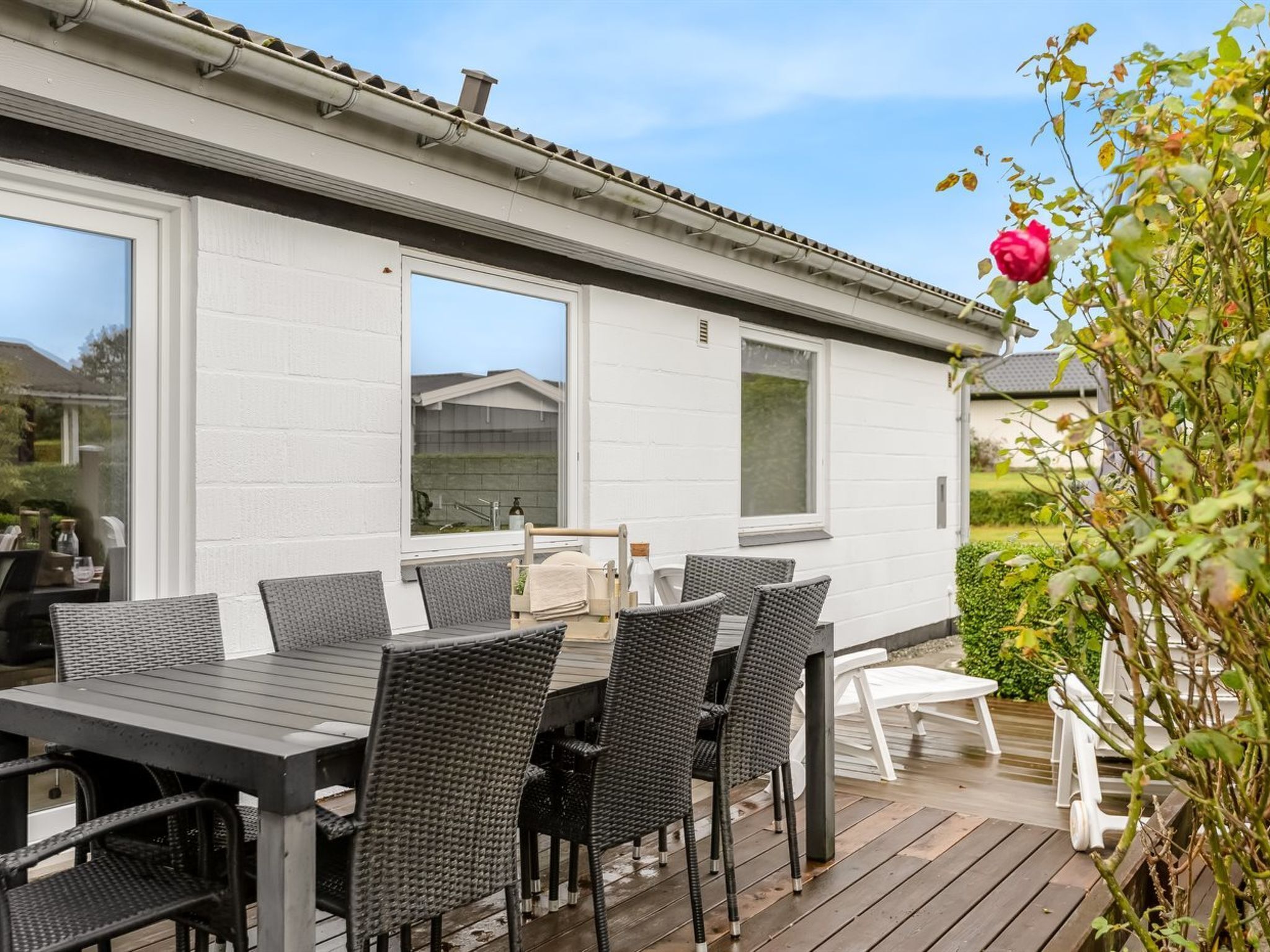 Een terras buiten met een eettafel en stoelen, een wit gebouw op de achtergrond en een rozenstruik in de voorgrond.