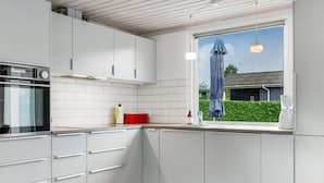 Cocina moderna con armarios blancos, horno empotrado y ventana con vista a una casa y vegetación.