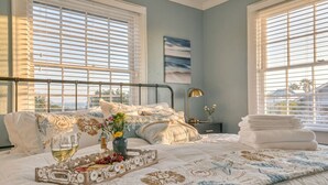 A bedroom with a bed, a bedside table with a lamp, a window with blinds, and a painting on the wall.