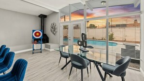 Ein modernes Zimmer mit einem Glastisch, blauen Stühlen, einem Zielscheibe an der Wand und Blick auf einen Swimmingpool.