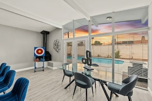 A modern room with a glass table, blue chairs, a target on the wall, and a view of a swimming pool. - Private home with pool! Sleeps 16 people (Las Vegas)