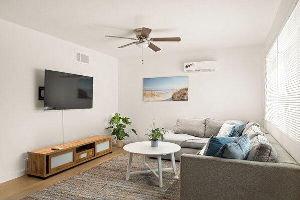 A modern living room with a flat-screen TV, ceiling fan, and a painting of a beach.