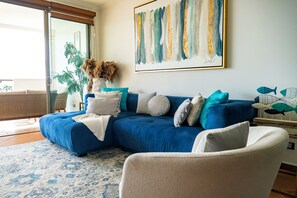 A living room with a blue sectional sofa, a white armchair, a large abstract painting, and a view of the outdoors through a sliding glass door.