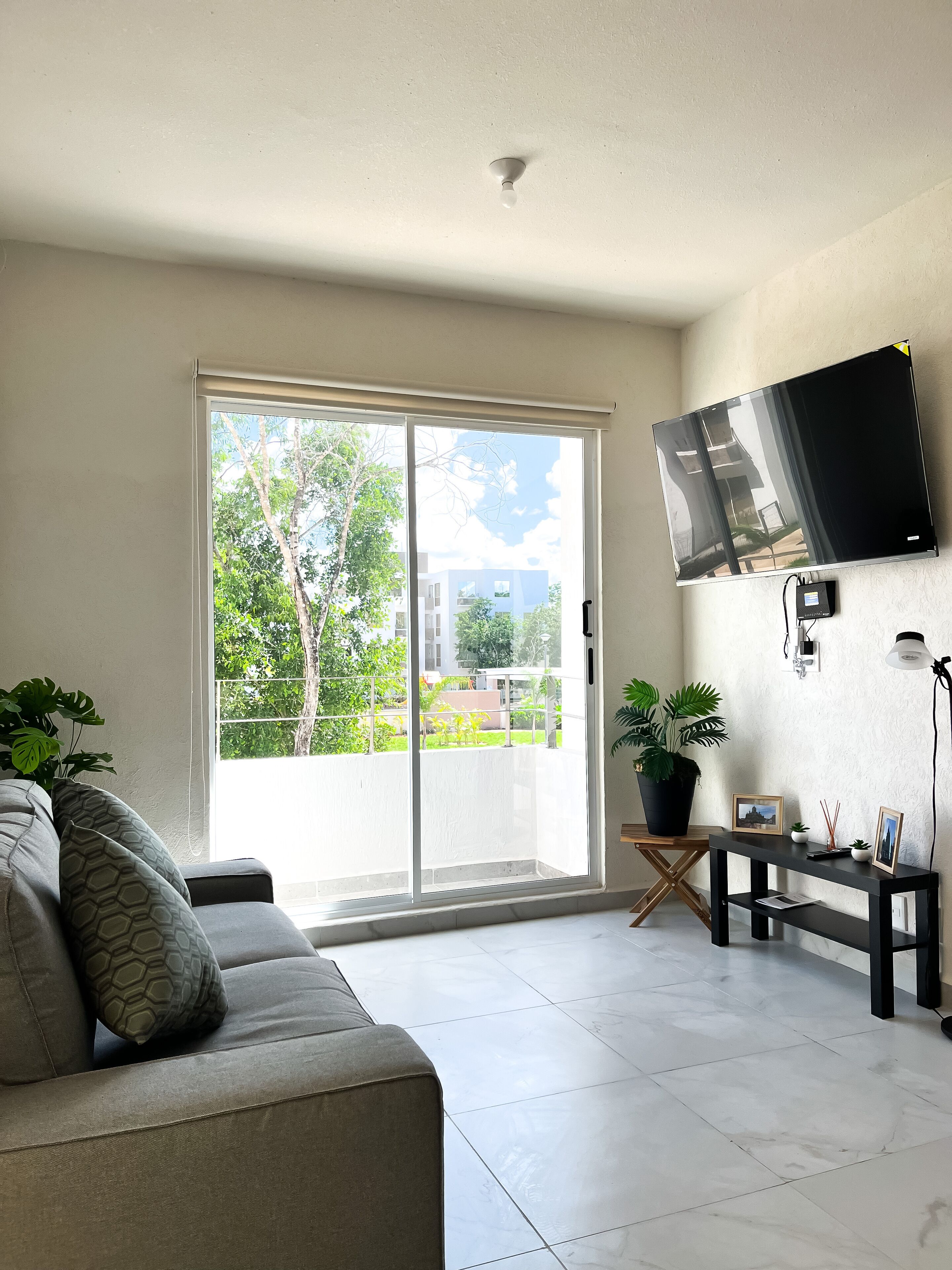 A modern living room with a sliding glass door leading to a balcony, a mounted flat-screen TV, a small table with decorative items, and a grey sofa with patterned cushions.