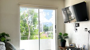A modern living room with a sliding glass door leading to a balcony, a mounted flat-screen TV, a small table with decorative items, and a grey sofa with patterned cushions.