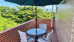 A balcony with a round table and two chairs, a green umbrella, and a view of lush greenery. - Charming 3-bedroom Apartment Near Puerto Plata's Malecon (Puerto Plata)