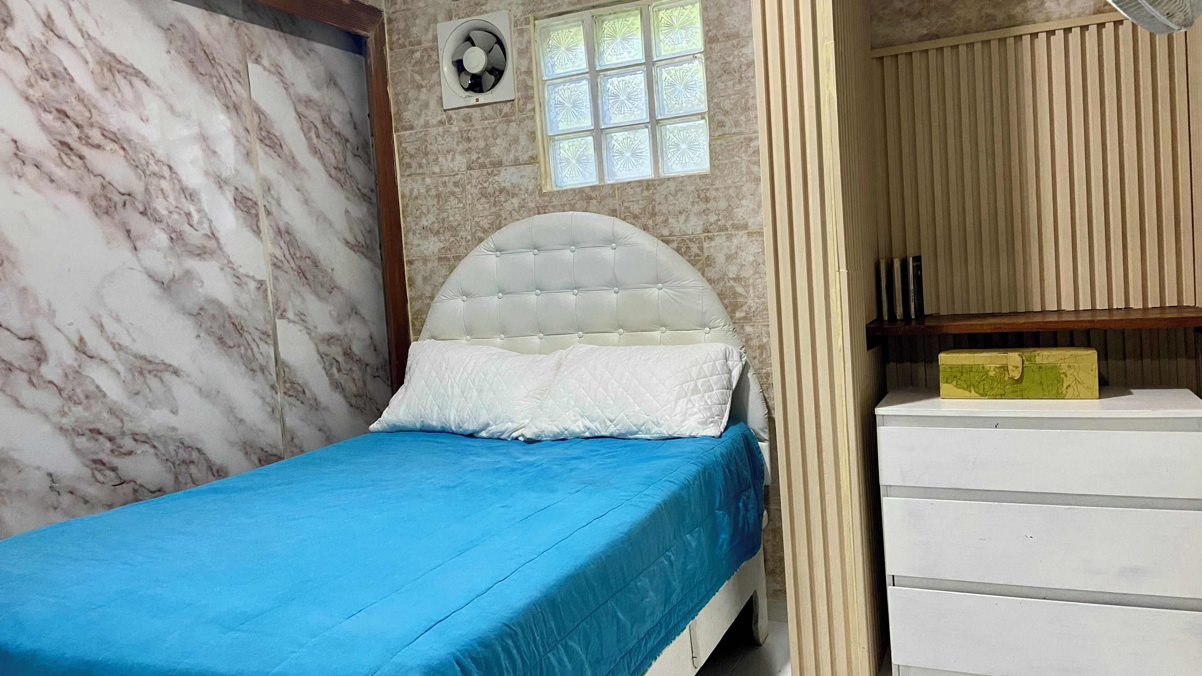 Une pièce comprenant un lit, une fenêtre ornée de vitraux, un ventilateur et une armoire en bois.