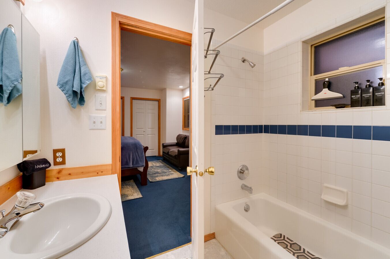 A bathroom with a bathtub, a sink, and a mirror. There is a door leading to a bedroom with a bed and a chair.