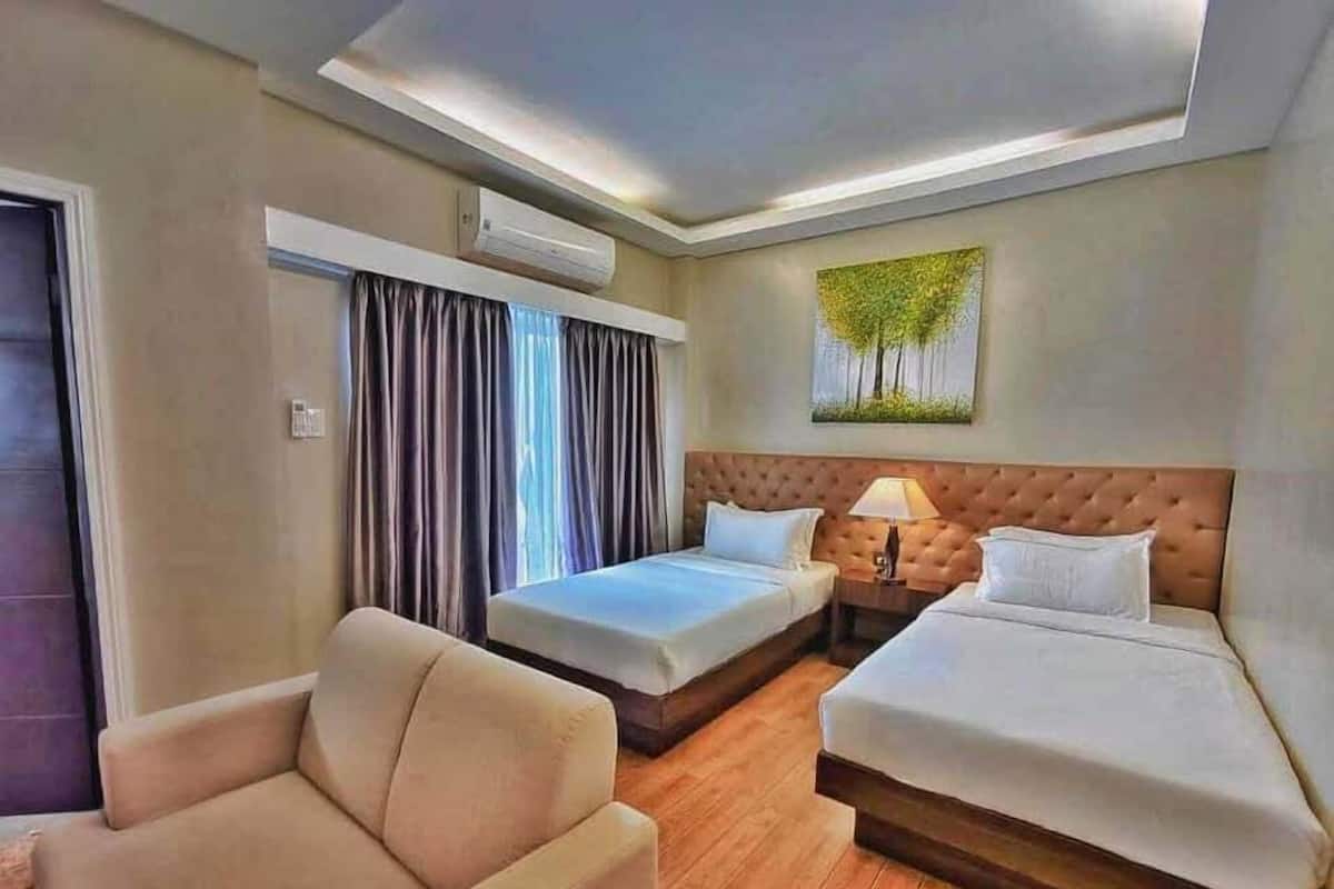 A hotel room with two beds, a sofa, a window with curtains, an air conditioning unit, and a painting on the wall.