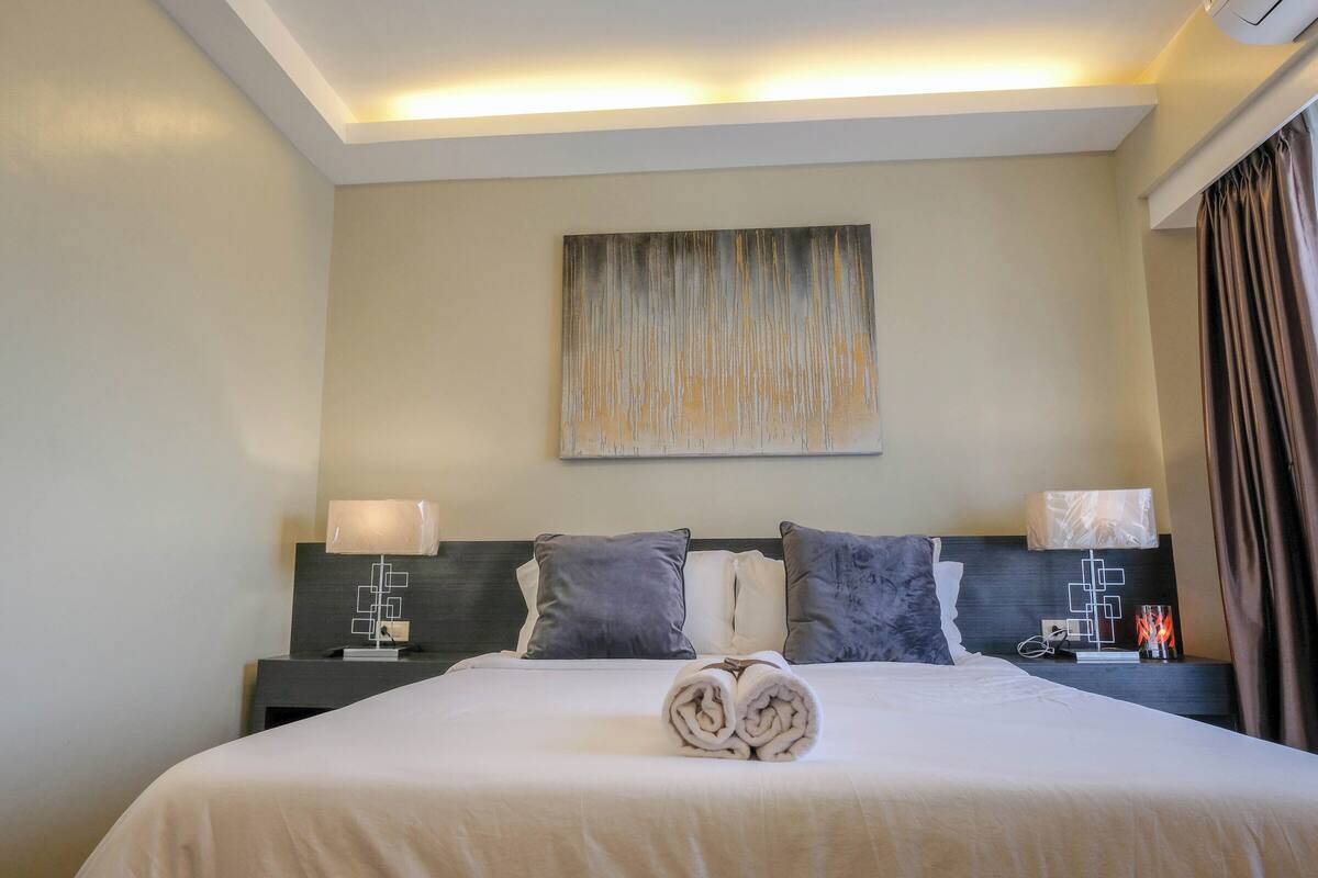A neatly made bed with a rolled towel in the center, a wall-mounted abstract painting, and a bedside table with a lamp and a small decorative item.