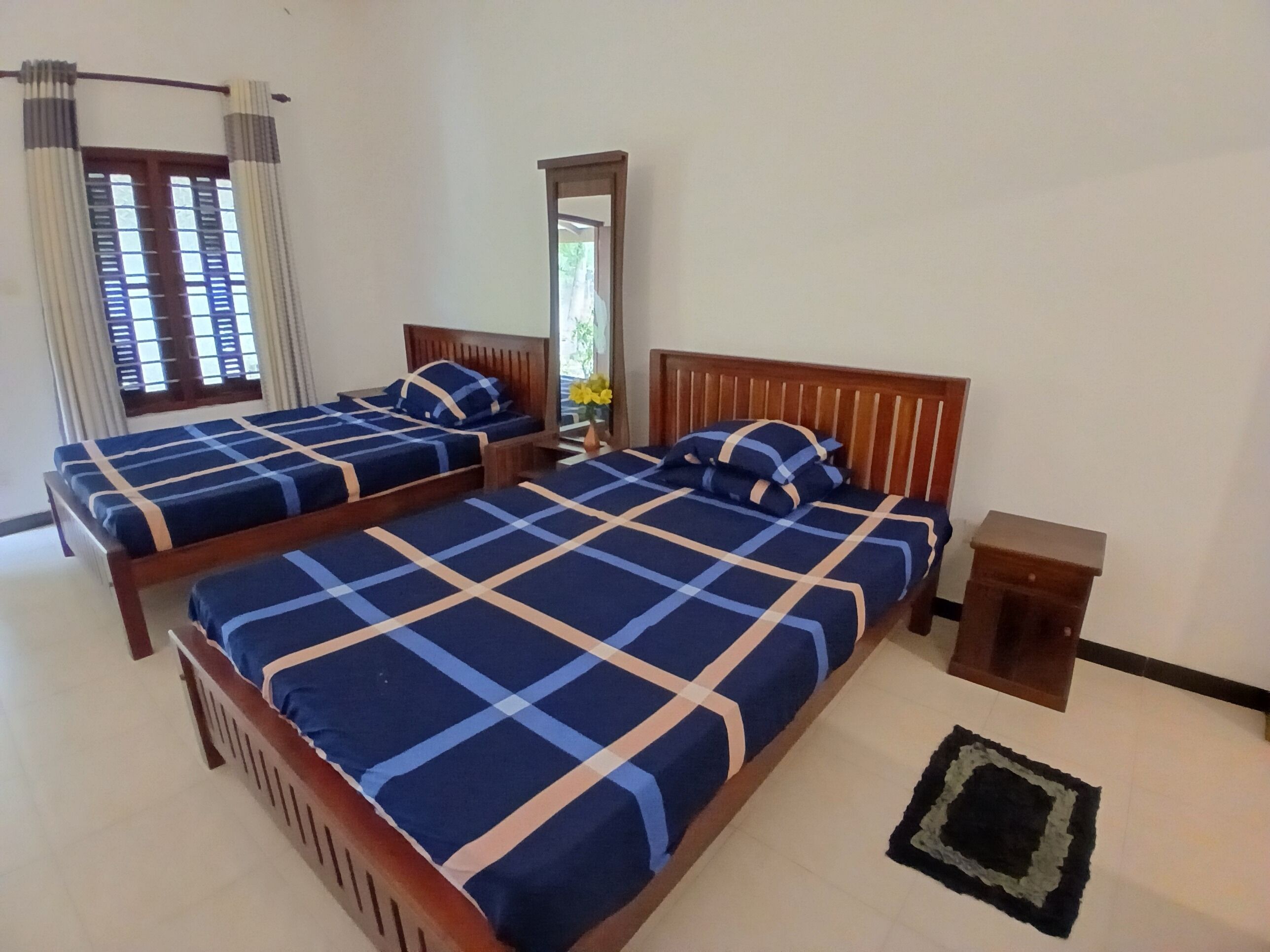 A room with two beds, each with a wooden headboard and blue bedding. There is a small wooden nightstand and a black rug on the floor.