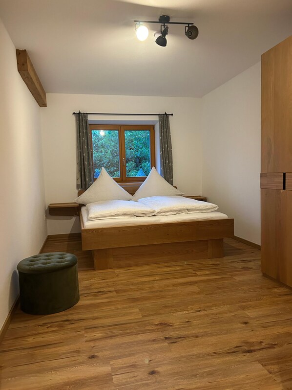 Une chambre avec un lit en bois, une fenêtre avec des rideaux, un petit tabouret et un luminaire au plafond.